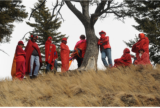 At the top of a small hill with dried, tall grasses in the foreground, nine people sit and stand surrounding a tree with branches. The people are all wearing red capes or wrapped in red fabric, some are wearing red masks or red hats. The group are strong looking, arms crossed, holding strong postures and looking down to the camera below.