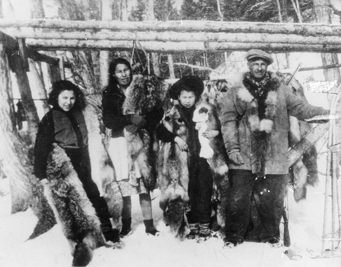 The black-and-white photograph depicts four individuals standing outdoors in a snowy forest setting. They are arranged in a line, with three people holding fur pelts. The individuals are dressed in winter clothing, including jackets and boots. The backdrop consists of snow-covered ground and barren trees, suggesting a cold environment. The person on the far right (Felix Beaver) wears a cap and a thick, fur-lined coat. A wooden structure made from logs is visible behind them.