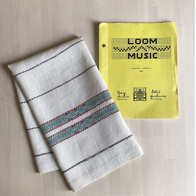 Photo: a small white towel with blue-grey line and two thicker turquoise lines on a table next to the yellow Loom Music book. 