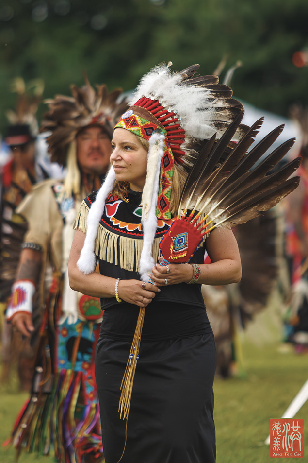 Colour photograph. A woman in Indigenous regalia is walking towards the camera, looking to the left. She is wearing a feathered headdress with a beaded fringe and long fur tassels, and is holding a large eagle feather fan. She is wearing a black dress fringed with buckskin and decorated with colourful beaded patterns around the shoulders. A group of Indigenous Dopeople wearing regalia are walking behind her in the background, out of focus.