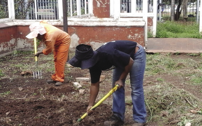 Colour photograph: Photo of two women in hats and jeans or coveralls hoeing the ground outside a house.