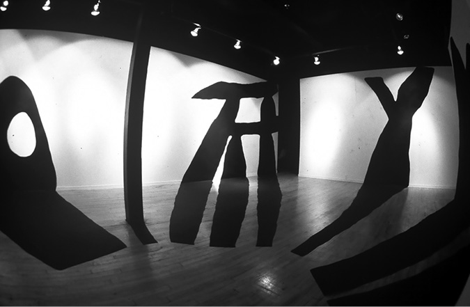 Black and white photograph: The Studio Gallery Nine gallery space with large, abstract, shadow-like cut outs laid across the floor and adhered the walls. The shadow on the back wall, in the right side of the image, looks like the letter Y.