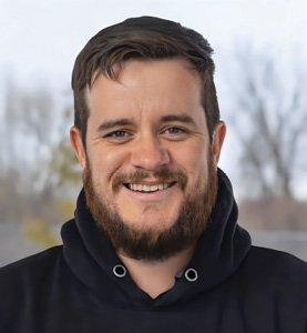 Colour photograph: Portrait of a smiling Indigenous man taken from the shoulders up. He has a beard and wears a black hoodie. He is standing outside with trees in the background.