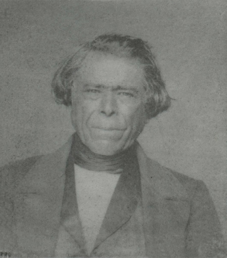 Black and white historical photograph. Head and shoulders portrait of an Indigenous man with black and mid-length hair covering his ears. He is wearing a suit jacket, waistcoat, and cravat and is looking into the camera.