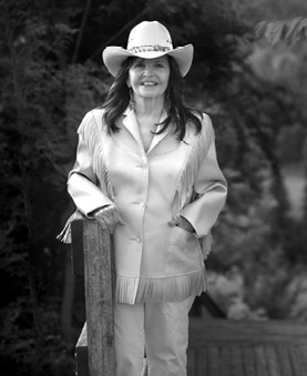 Black and white photograph: A woman stands outdoors wearing a cowboy hat, a fringed jacket, and matching pants. She is resting her hands in her pockets and smiling at the camera. Trees and greenery can be seen in the background.