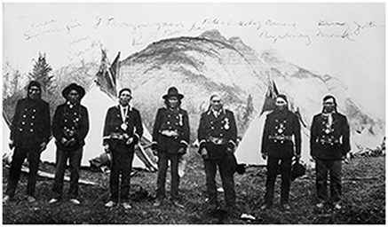 Photograph of James Swampy, Hector Crawler, Jonas, Two Young Man, John Bearspaw, Peter Wesley, Amos Big Stoney, John Mark. Standing In front of the tent, with the mountains as a backdrop.