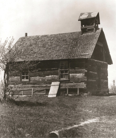 Black and white historical photograph. A log schoolhouse, with a shingled roof. There are two windows, a door on the side, a cellar door, and a wooden bell tower. The building is in a field with a few small trees.