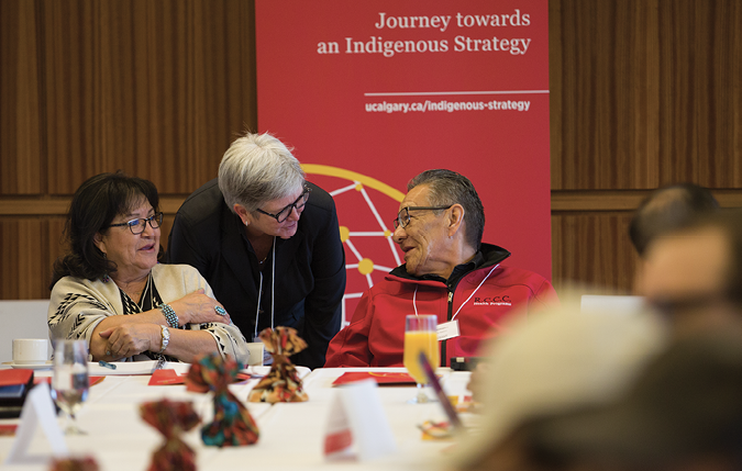 Colour Photograph: Dru Marshall talking to two Elders (Evelyn Goodstriker and Calvin Williams) in front of Journey towards an Indigenous strategy banner.