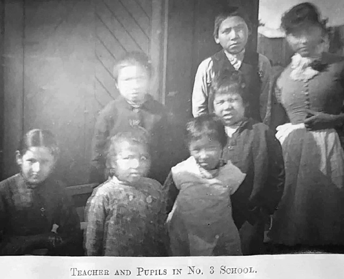 Black and white photograph: Somewhat blurry older candid photograph from the 1800s of six male and female Indigenous students and their teacher in front of a small building (likely a day school).  The students are dressed in a variety of casual clothing. The caption under the photo reads “Teacher and Pupils in No. 3 School.”