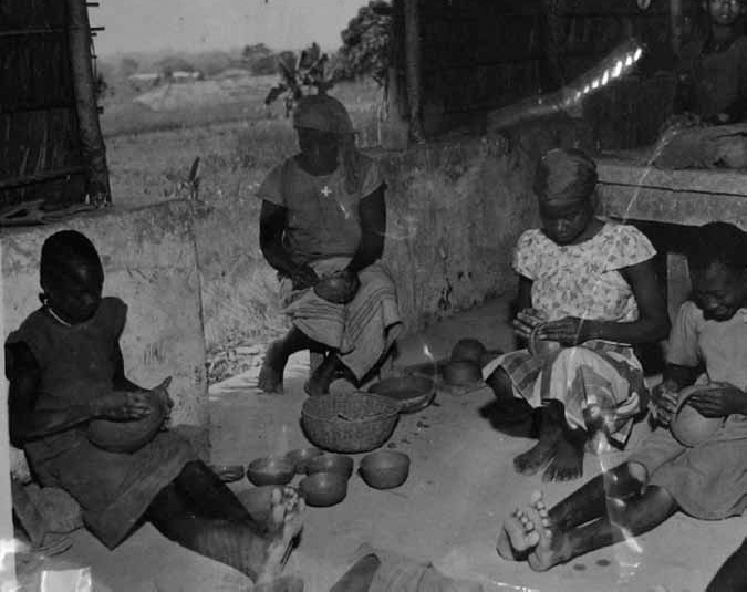 A black and white photograph shows tribal women making pottery products. Each is holding different sizes of pottery. The finished pottery is arranged beside them.