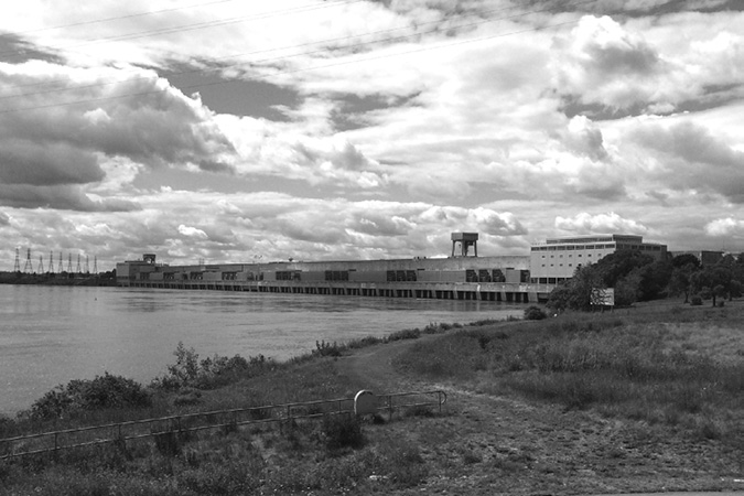A photograph of the Moses-Saunders powerhouse, a long, low building situated on Lake St. Lawrence. The building is in the centre of the photograph under a bright, cloudy sky, with Lake St. Lawrence visible on the left.