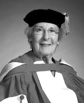Black and white photograph: A portrait of an elderly woman wearing academic regalia, including a gown and a hat with a tassel. She has short hair, glasses, and a gentle smile.