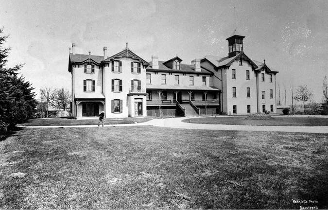Black and white photograph: Second set of Mohawk Institute buildings in the 1890s.  There is a large two-storey brick building in the middle, and significant additions built on each side of the school. There is a porch across the first level of the main building and a set of stairs leading to the front door. There is a circular drive located in front, as well as grassed areas.  A man is raking the grass. 