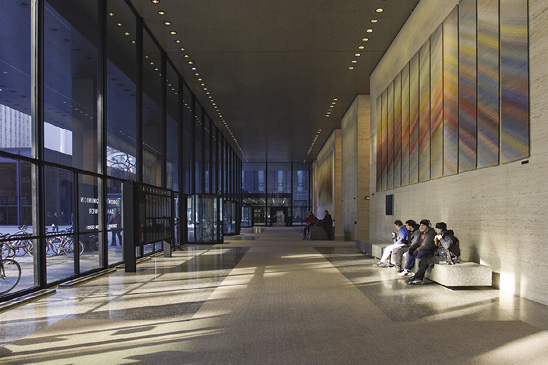 Photo: a long hallway inside a wall of glass. Light shines through onto four people sitting on a bench beneath an artwork made up of multiple vertical panels.