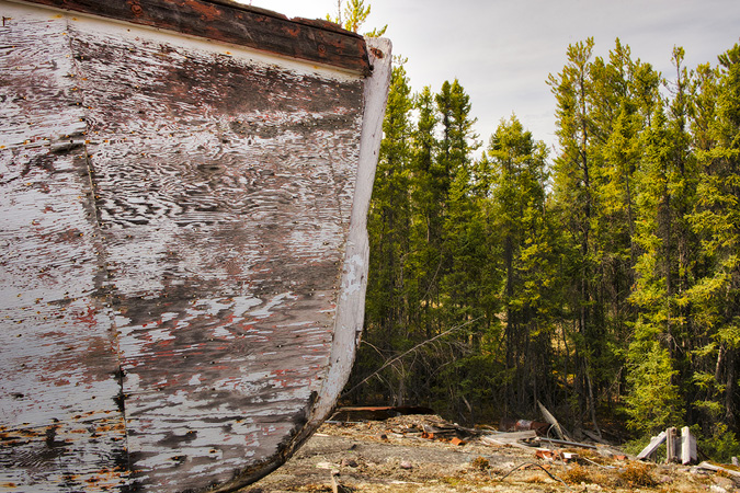 The weathered prow of a large wooden boat, stranded in a thick coniferous forest, dominates the left-hand side of this image; the forest takes up the right-hand side of the image, which was gathered by the side of a grid road near Uranium City, Saskatchewan.