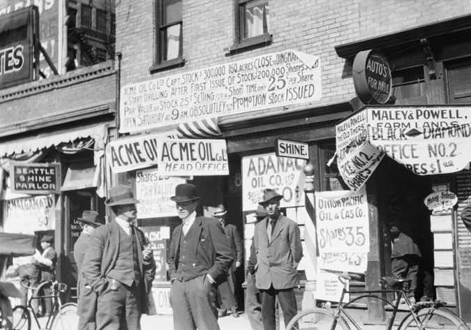 Figure 5-5 “Investors waiting to buy oil stocks, Calgary, Alberta.” Black and white image taken outside Acme Oil Company’s head office—216, 8th Avenue SW. The location is less than a three-minute walk from the Canadian Pacific Railway station, and illustrates the various ways that promoters attempted to capture people’s attention to buy oil stock. In this image, hand-made signs are visible for Amalgamated, Acme Oil, Adanac Oil Company, Black Diamond #2, and Union Pacific Oil and Gas Company. In the foreground are three men dressed in suits and ties as another stands behind them in the door of Acme Oil. Two bicycles are parked along the sidewalk.
