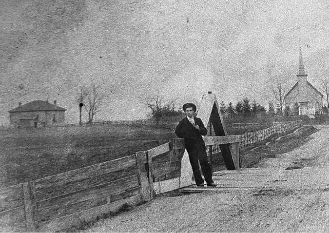 Black and white photograph: Older photograph shows a man outdoors leaning on a fence beside a long driveway that leads to a wooden chapel.  In the left background, there is a large brick building which may be the Mohawk Institute.
