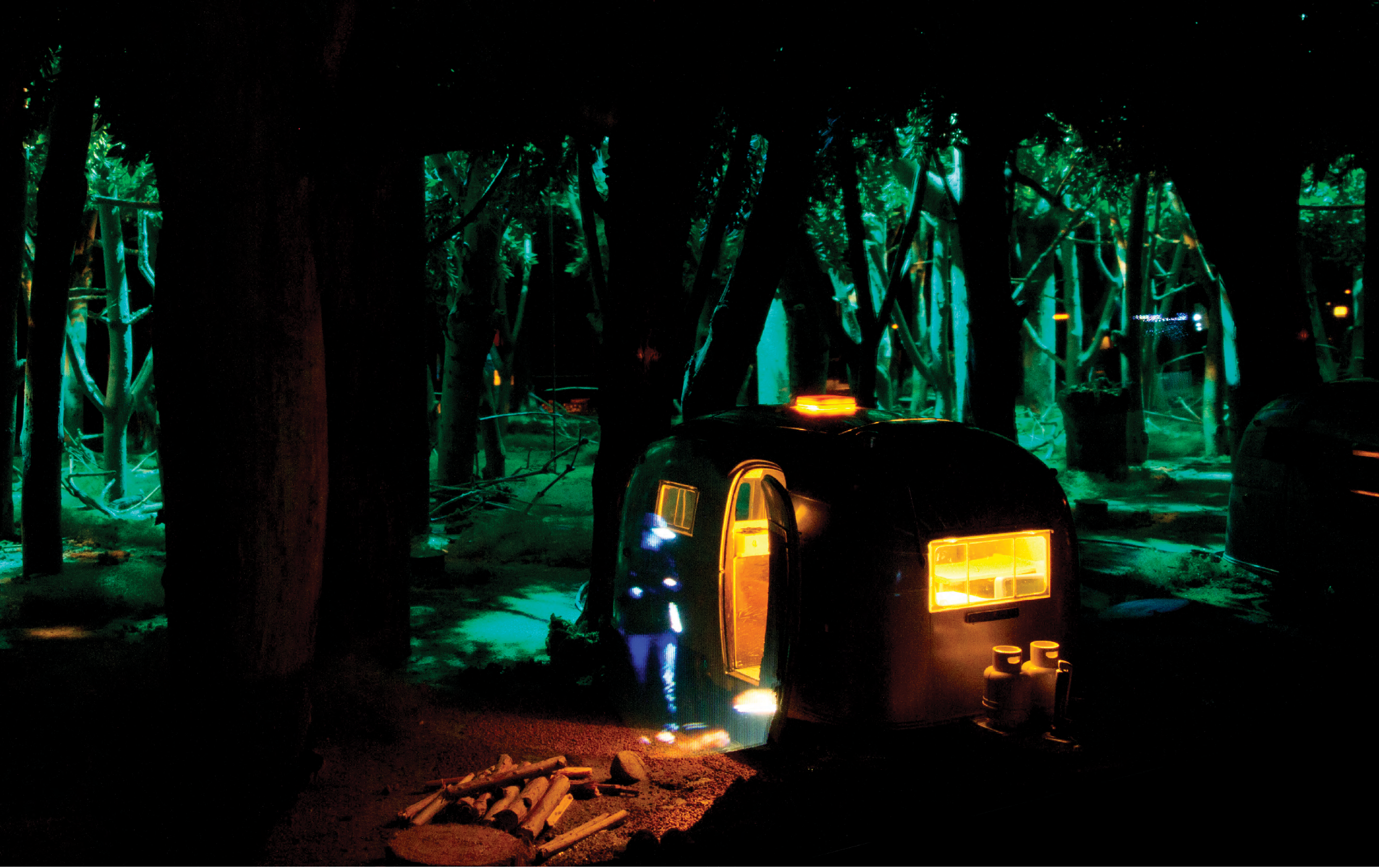 Colour photograph: A diorama with projections, of a figure standing in front of the door of a small travel trailer, in the woods at night.