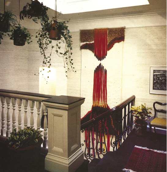 Photo: on the landing at the top of stairs, a large hanging that is brown along the top and mostly white but with long ropes of red down the middle. Four plants hang from the ceiling. 