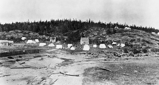 Black and white photograph: An encampment comprising three log homes and several white tents and tipis. The ground is dry, covered only by rocks and shrubs. In the centre is a pile of long, wooden posts. To the left is a wooden post fence. Two long canoes lie in the foreground, one lying upright, and one positioned on its side. In the distance, people are walking amongst the tents. Behind the encampment is the forest.
