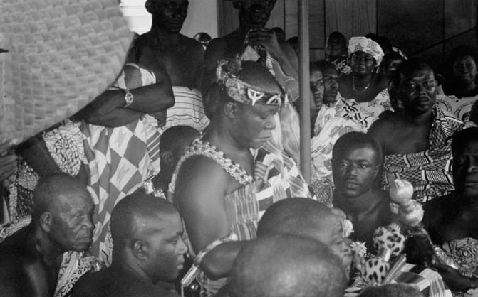 A black and white photograph of a group of African people standing and some sitting in the middle, all wearing African dresses.