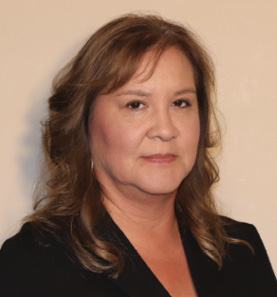 Colour Photograph. Portrait of an Indigenous woman taken from the shoulders up. She has shoulder-length hair and is wearing a black blazer. 