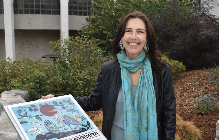 Colour photograph. An Indigenous woman is standing in a garden in front of a modern building, with her hand on an interpretive plaque. She is smiling and has long brown hair. She is wearing large earrings, a long turquoise scarf, and a dark leather jacket. The plaque features her Woodlands Style painting of various local birds and plants.