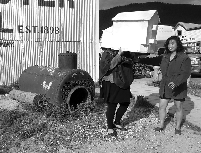 Black and white photograph, two women stand beside a wooden boardwalk, the woman on the left holds a small white object shaped like a house over her head while the woman on the right holds her shoulder, a disused boiler and corrugated iron building stand beside the women, and a view of wooden buildings and cars is visible in the background.