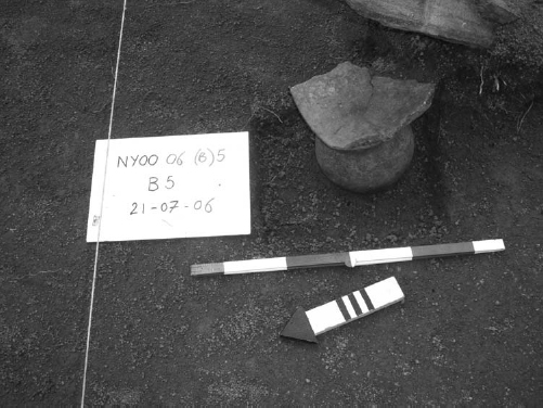 A short, spherical jar with a potsherd for a lid. Text on a card placed on the site reads, NYOO 06 (B)5. B 5, 21-07-06.
