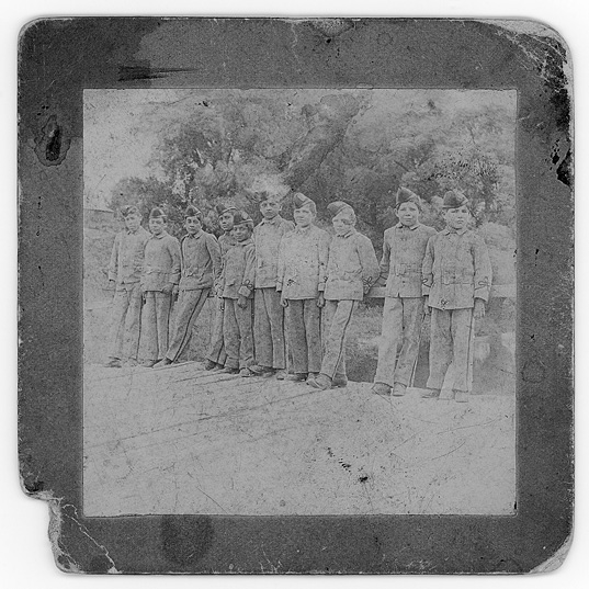 Black and white photograph of a photograph or slide: Older photograph from the 1890s that is somewhat blurry.  There is a tattered edge. Ten Indigenous male student cadets are casually posed in a row wearing cadet uniforms and caps.