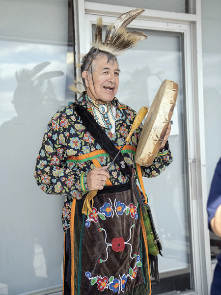 Colour photograph. A man is standing in colourful Indigenous dress in front of a glass wall. He is singing and playing a hand drum. He wears a feathered head piece, a colourful floral ribbon shirt, a beaded medallion with bone bead (hairpipe) breastplate, and a black apron decorated with beaded floral patterns.